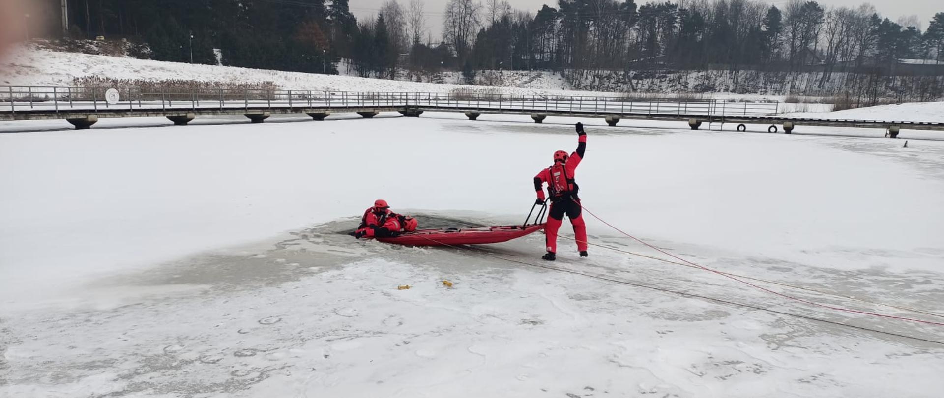 Ratownik w czerwonym kombinezonie znajduje się częściowo zanurzony w przeręblu, trzymając się czerwonych sani ratowniczych. Drugi ratownik stoi na lodzie, ciągnąc sanie za pomocą liny i unosząc rękę – prawdopodobnie w celu komunikacji. W tle widoczny jest most oraz las, a całość rozgrywa się w śnieżnym, zimowym krajobrazie. Zdjęcie ukazuje dynamiczny moment akcji ratunkowej na lodzie.