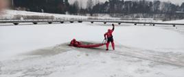 Ratownik w czerwonym kombinezonie znajduje się częściowo zanurzony w przeręblu, trzymając się czerwonych sani ratowniczych. Drugi ratownik stoi na lodzie, ciągnąc sanie za pomocą liny i unosząc rękę – prawdopodobnie w celu komunikacji. W tle widoczny jest most oraz las, a całość rozgrywa się w śnieżnym, zimowym krajobrazie. Zdjęcie ukazuje dynamiczny moment akcji ratunkowej na lodzie.