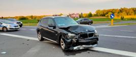 Zdjęcie przedstawia uszkodzony czarny samochód marki BMW, z którego wyciekł płyn eksploatacyjny. Na drugim planie znajdują się inne samochody oraz policyjny radiowóz.