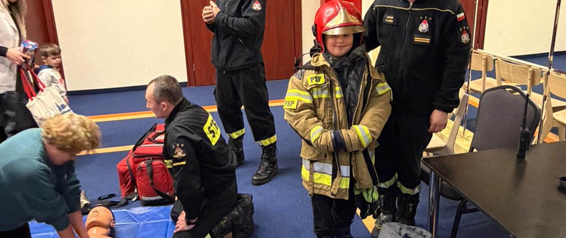 W sali z drewnianymi drzwiami i panelami ściennymi kobieta wykonuje uciski klatki piersiowej na fantomie leżącym na niebieskiej macie. Obok niej klęczy strażak w czarnym mundurze, instruując ją. W tle obecni są inni strażacy w czarnych mundurach z żółtymi pasami. Dziecko w stroju strażackim z napisem „OSP” stoi obok dorosłego w mundurze. Wokół rozstawione są krzesła, a atmosfera wskazuje na szkolenie z zakresu pierwszej pomocy