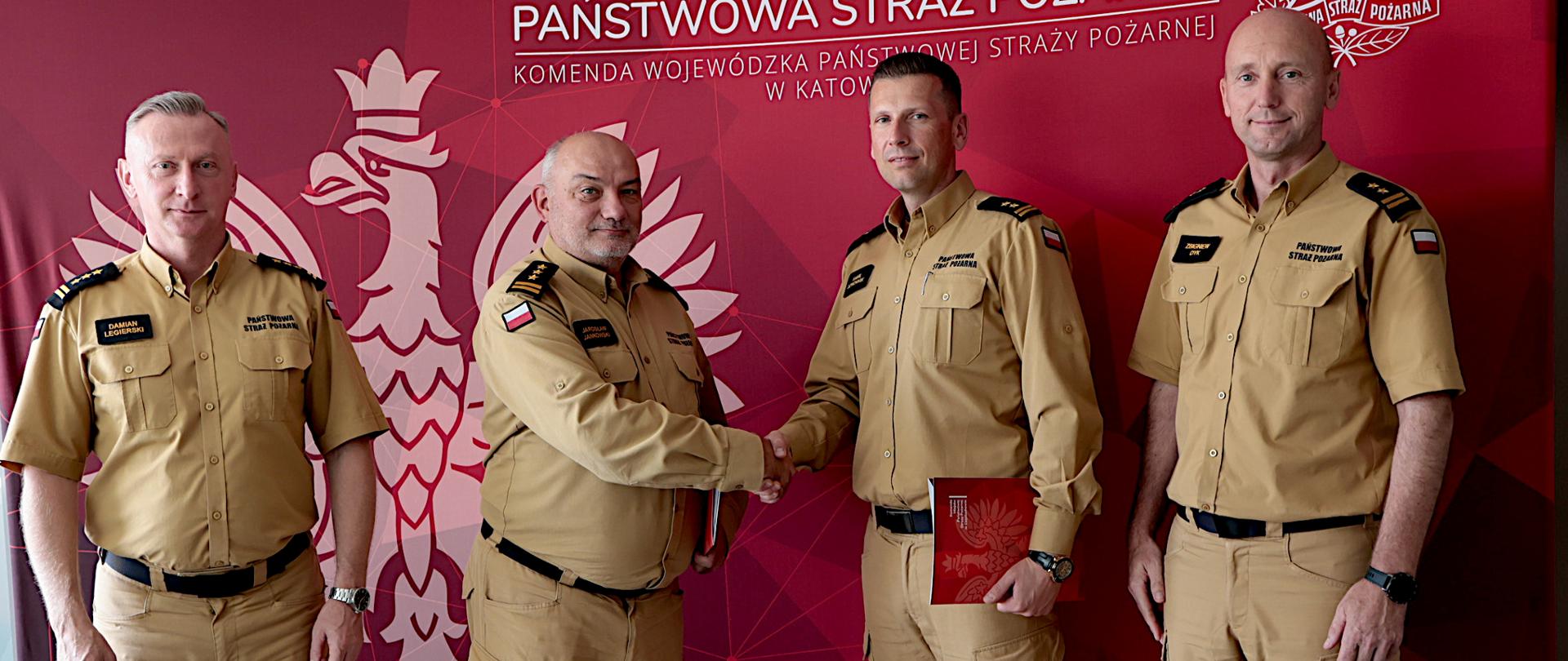 Na zdjęciu stoją czterej strażacy Państwowej Straży Pożarnej w piaskowych mundurach, od lewej strony śląski komendant wojewódzki Państwowej Straży Pożarnej, komendant Centralnej Szkoły Państwowej Straży Pożarnej w Częstochowie, komendant miejski Państwowej Straży Pożarnej w Częstochowie oraz zastępca śląskiego komendanta wojewódzkiego Państwowej Straży Pożarnej. Stoją oni na tle czerwonej ścianki z dużym godłem i napisem Komendy Wojewódzkiej Państwowej Straży Pożarnej w Katowicach