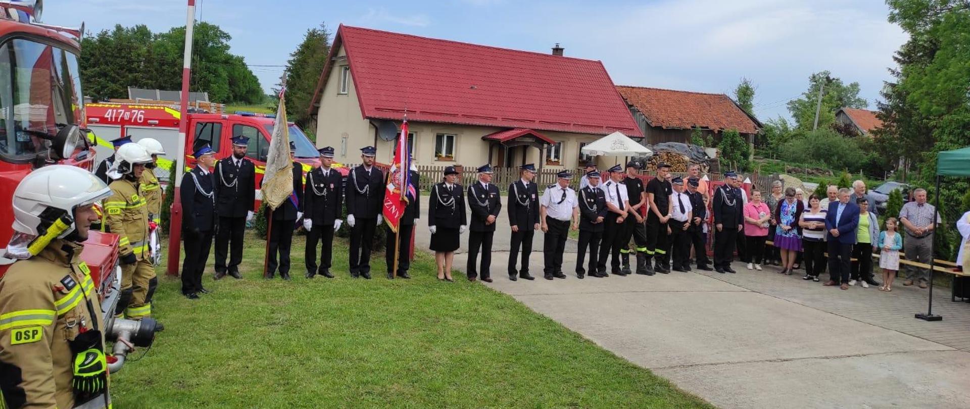 Plac przed remizą. Z lewej strony strażacy w mundurach bojowych i białych hełmach. Dalej na wprost strażacy ze sztandarami w mundurach galowych, obok kolejni strażacy i zaproszeni goście.