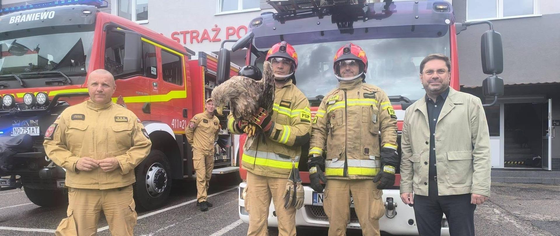 Przed remizą strażacką stoi pięciu mężczyzn. Trzech strażaków jest na pierwszym planie, jeden trzyma dużego ptaka drapieżnego; za nimi widać czerwone wozy strażackie z drabiną.