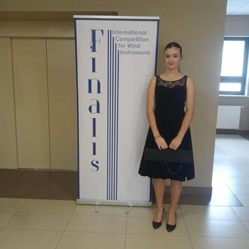 Zosia Zając stojąca z fletem w futerale na korytarzu przy plakacie konkursu Finalis.