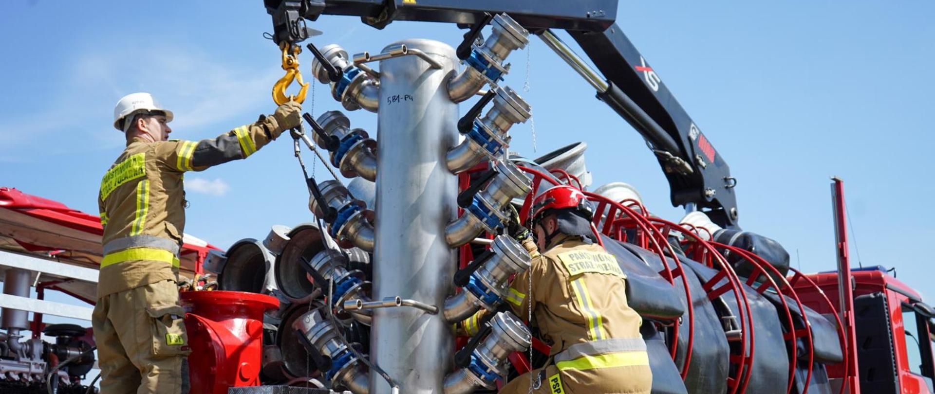 Operator dźwigu wraz ze strażakiem montują kolektor wodny z wieloma przyłączami na platformie pojazdu.