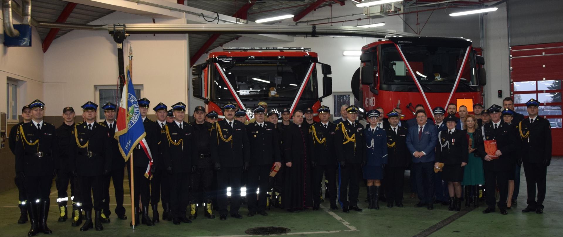 Przed nowymi pojazdami pożarniczymi ozdobionym biało-czerwonymi wstęgami stoją uczestnicy uroczystej zbiórki funkcjonariusze, przedstawiciele firm oraz instytucji.