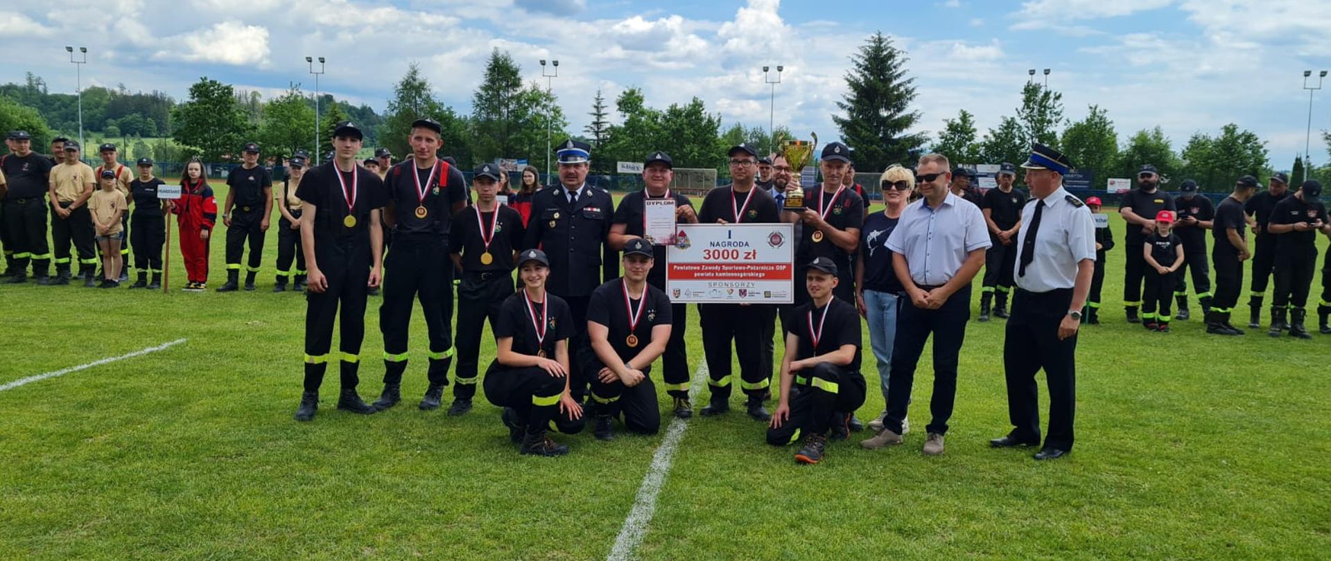 Drużyna startująca w sporcie pożarniczym, jeden z zawodników trzyma przed sobą główną nagrodę czek na trzy tysiące złotych. W tle pozostałe osoby startujące w zawodach ustawione w szeregu. Zdjęcie wykonane na murawie boiska sportowego.