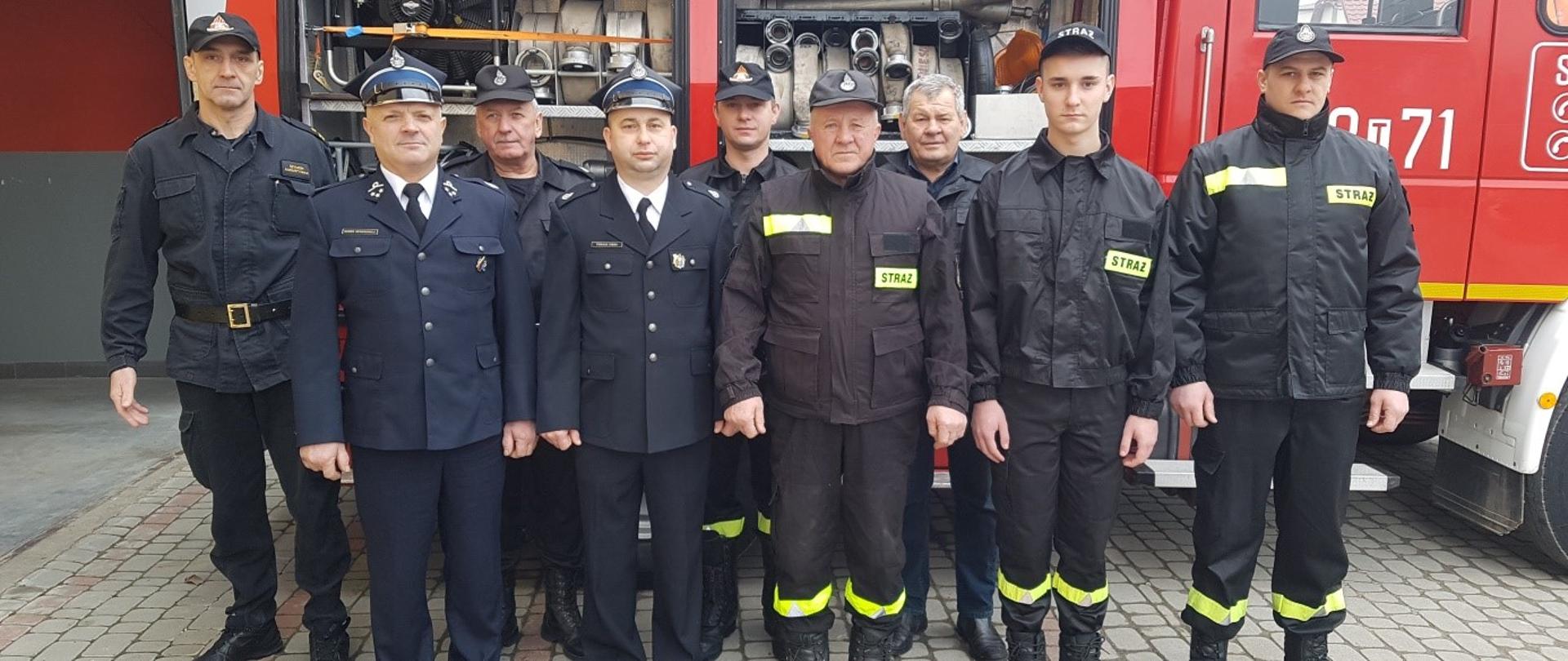 strażacy ochotnicy oraz oficer PSP stoją na tle samochodu gaśniczego. Samochód czerwony z podniesionymi skrytkami na sprzęt (węże ułożone na półkach).