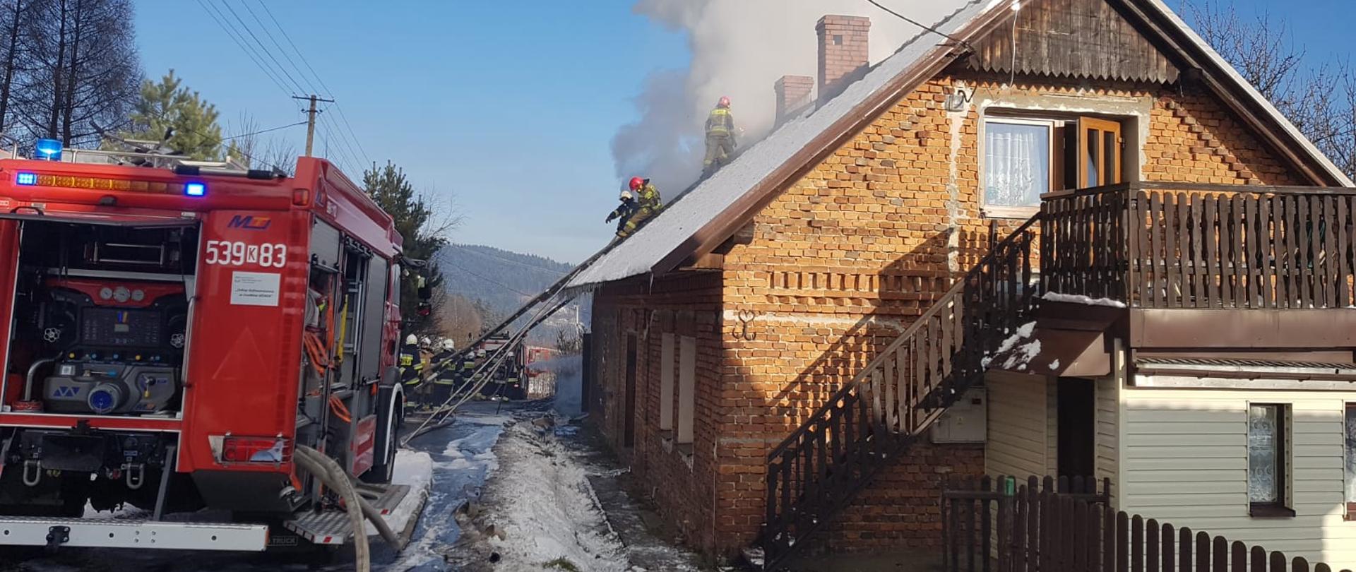 Zdjęcie przedstawia palący się budynek gospodarczy ze strony budynku mieszkalnego, z budynku wydobywa się dużo dymu. Strażacy na dachu walczą z ogniem, próbując uratować część mieszkalną. Na zdjęciu widać samochód pożarniczy podający wodę do pożaru.