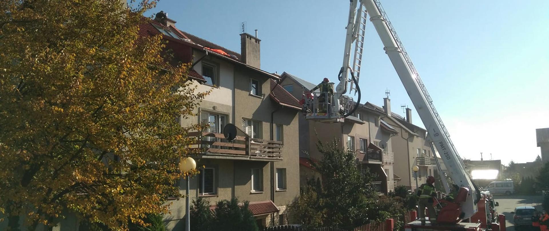 Zdjęcie przedstawia strażaków w koszu podnośnika hydraulicznego, podejmujących ewakuację spadochroniarza, który wylądował na dachu budynku.