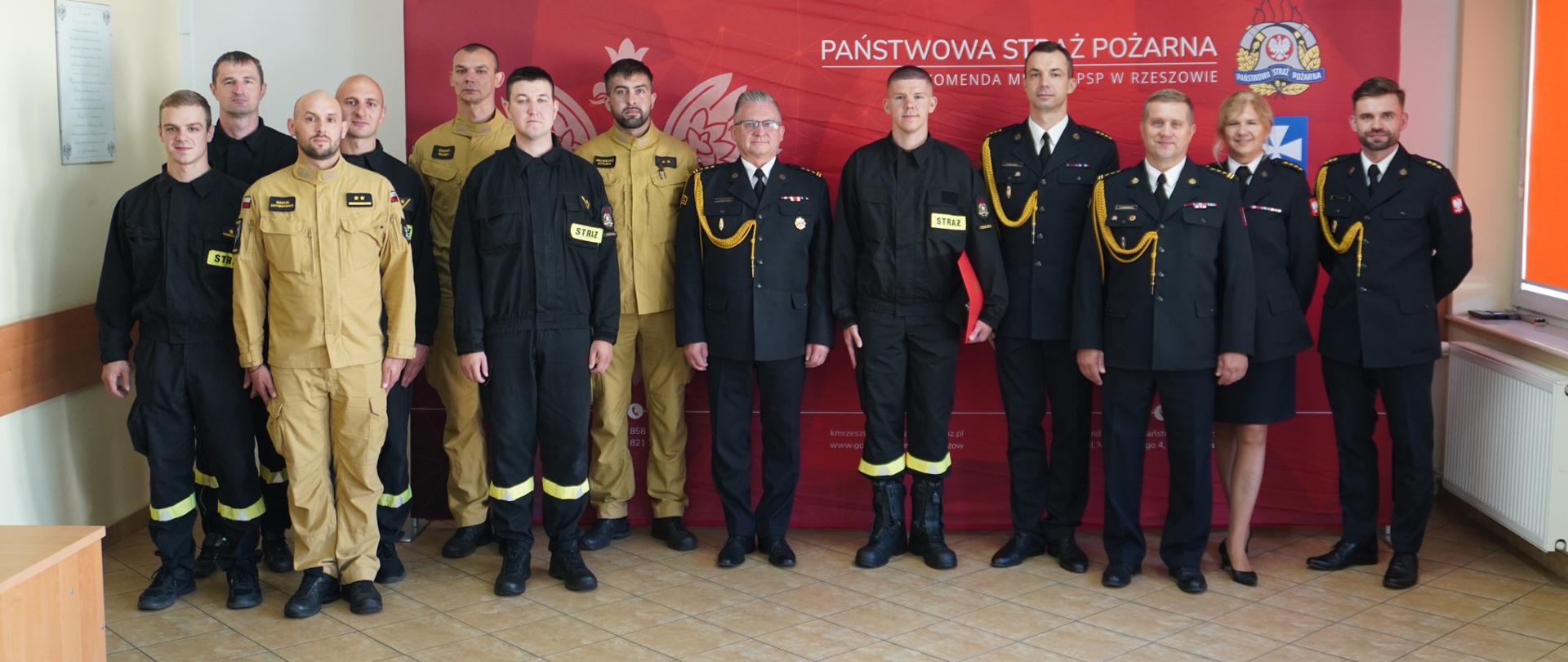 Wspólne zdjęcie grupowe strażaków PSP w Rzeszowie po zakończeniu uroczystości. Na tle czerwonej ścianki z godłem Polski i logotypem Państwowej Straży Pożarnej stoi kilkanaście osób w dwóch rzędach. W pierwszym rzędzie znajdują się funkcjonariusze w czarnych i piaskowych ubraniach bojowych, z naszywkami „STRAŻ” na kurtkach. Pośrodku stoją oficerowie w mundurach galowych z odznaczeniami i złotymi sznurami, a z prawej strony także kobieta w galowym mundurze. Drugi rząd tworzą strażacy w bojowych ubraniach roboczych. Grupa stoi równo w szeregu, patrząc w stronę aparatu fotograficznego.