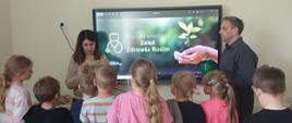 Grupa dzieci uczestniczy w zajęciach edukacyjnych o zdrowiu roślin. Kobieta pokazuje dzieciom materiał roślinny, a obok stoi mężczyzna. W tle na ekranie widoczny jest napis „Międzynarodowy Dzień Zdrowia Roślin” oraz grafika dłoni trzymających sadzonkę.