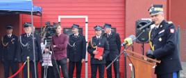 Strażak Państwowej Straży Pożarnej stoi przy mównicy i przemawia do mikrofonów obok stoją zaproszeni goście oraz przedstawiciele prasy.