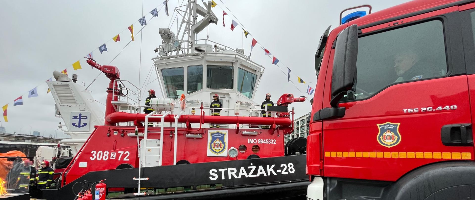 Statek Strażak 28, na nim strażacy Portowej Strazy Pożarnej w umundurowaniu, na zdjęciu znajduje się także bok samochodu ratowniczo gaśniczego Portowej Straży Pożarnej