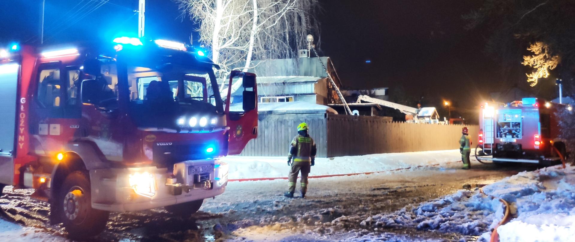 Zdjęcie przedstawia pojazdy pożarnicze oraz strażaków podczas działań ratowniczo-gaśniczych na tle budynku objętego pożarem