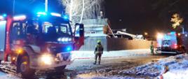 Zdjęcie przedstawia pojazdy pożarnicze oraz strażaków podczas działań ratowniczo-gaśniczych na tle budynku objętego pożarem