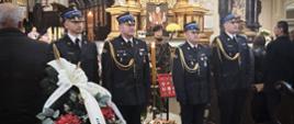 Strażacy w mundurach galowych stoją na baczność przed trumną śp. płk. poż. Stanisława Luli. Obok widoczny wieniec z biało-czarną wstęgą pożegnalną oraz medalami i odznaczeniami zasłużonego oficera.