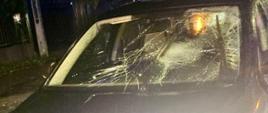 Ciemny samochód osobowy marki Skoda z widocznymi uszkodzeniami przedniej szyby, która jest popękana w wielu miejscach. Zdjęcie wykonano nocą, podczas opadów deszczu, na oświetlonej ulicy.