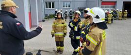 Na zdjęciu widać strażaka z Państwowej Straży Pożarnej oraz pięcioro druhów z OSP. Obok nich na macie sprzętowej leżą aparaty Ochrony Układu Oddechowego oraz piła do cięcia drewna. Funkcjonariusz z Państwowej Straży Pożarnej tłumaczy druhom zasady egzaminu obowiązujące na stanowisku ze znajomości sprzętu ratowniczo – gaśniczego. W tle widać grupę druhów z OSP czekających przed budynkiem Komendy.