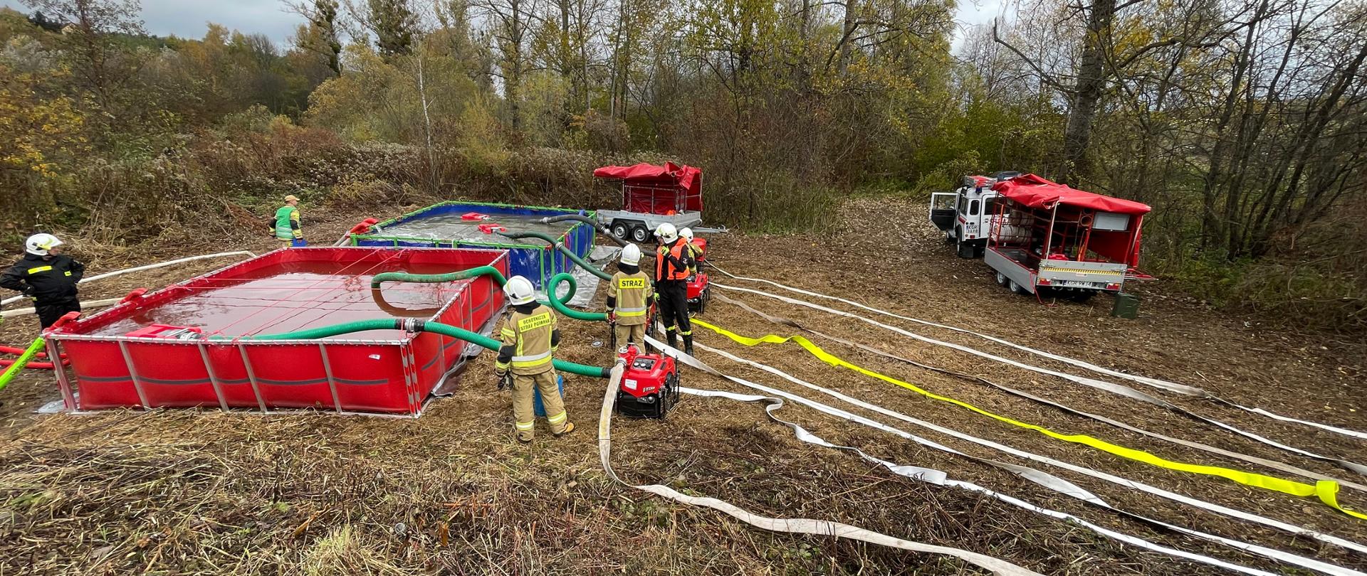 Na zdjęciu znajduje się stanowisko wodne przy rzece. Po lewej stronie ustawione są dwa zbiorniki brezentowe wypełnione wodą. Na środku zdjęcia znajduje się czterech strażaków przy motopompach pożarniczych. Od motopomp rozciągają się linie wężowe. W tle widać krzaki oraz drzewa. 