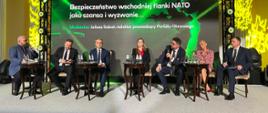Przed czarno-zieloną ścianką z napisem "Bezpieczeństwo wschodniej flanki NATO jako szansa i wyzwanie" na krzesłach siedzi siedem osób, trzeci od prawej wiceminister Szeptycki mówi do trzymanego w ręku mikrofonu.