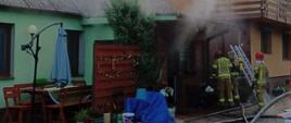 Zdjęcie przedstawia pożar kotłowni zlokalizowanej przy domu mieszkalnym. Z pomieszczenia kotłowni wydobywa się dym. Przed budynkiem widać strażaków oraz przystawioną do budynku drabinę. Na zdęciu widać również węże strażackie 