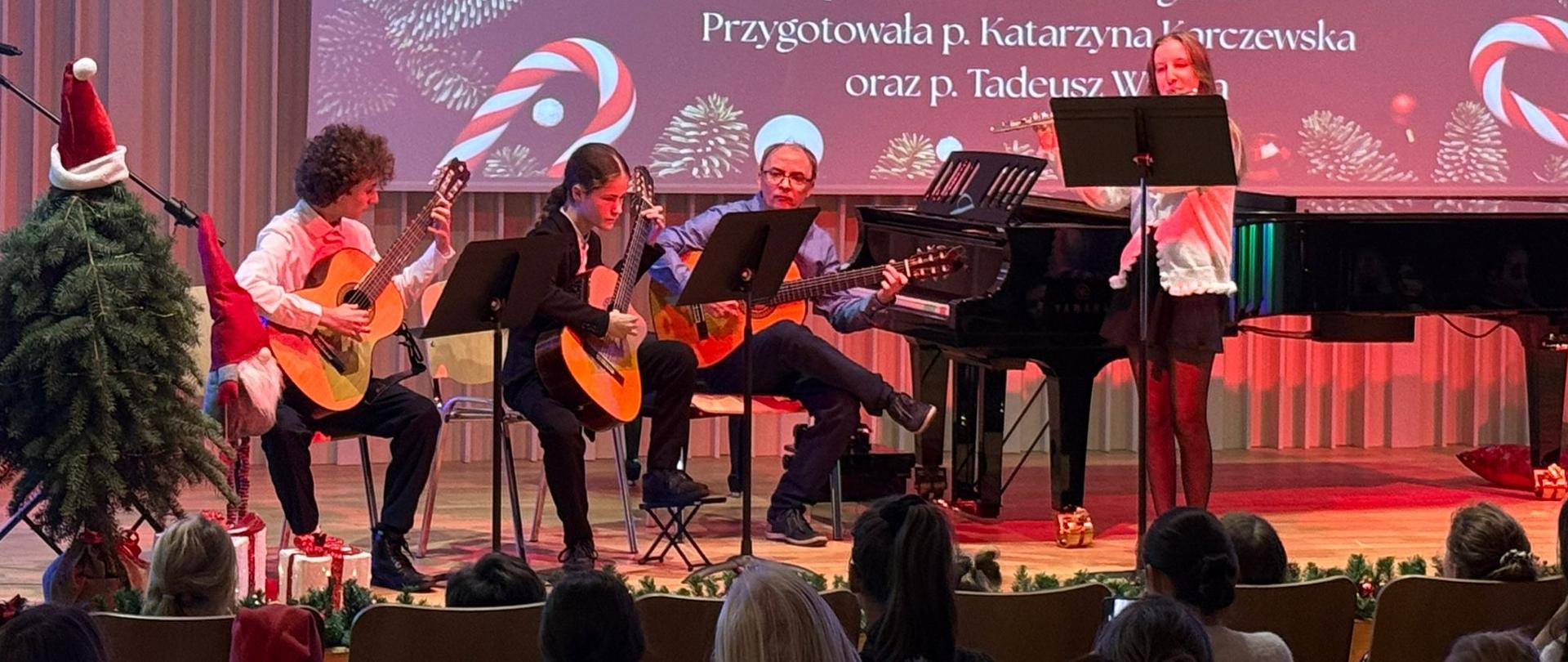 Na zdjęciu przedstawiony jest występ instrumentalno-wokalny podczas szkolnego koncertu świątecznego. Na scenie siedzi trzech uczniów grających na gitarach klasycznych, a obok nich stoi uczennica grająca na flecie przy pulpicie z nutami. W tle widoczny jest fortepian oraz ekran z bożonarodzeniową grafiką i napisem „Mizerna Cicha, Gdy śliczna Panna” wraz z informacją o wykonawcach. Scena udekorowana jest świątecznie – widać choinkę, skrzata oraz drobne dekoracje. Na pierwszym planie znajduje się publiczność siedząca na widowni. Całość oddaje uroczysty i świąteczny charakter koncertu.