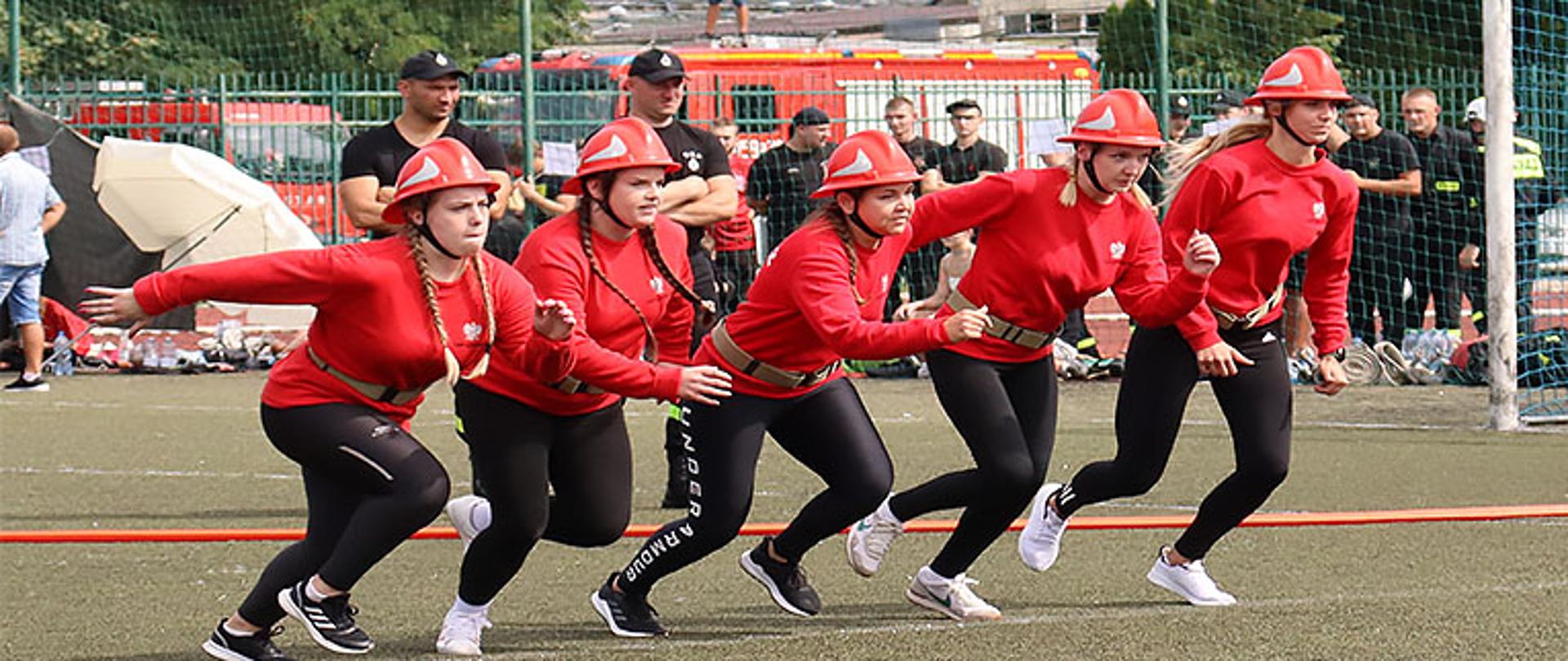 grupa pięciu zawodniczek podczas startu do bojówki strażackiej na murawie boiska, ubrane są w czerwone bluzy i czarne spodnie, na głowie mają czerwone hełmy, w tle zawodnicy oraz służby porządkowe 