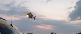 Zdjęcie helikoptera LPR na tle nieba. W dolnym rogu samochód ZRM w kolorze kanarkowym. W oddali drzewa. Zachmurzenie lekkie.