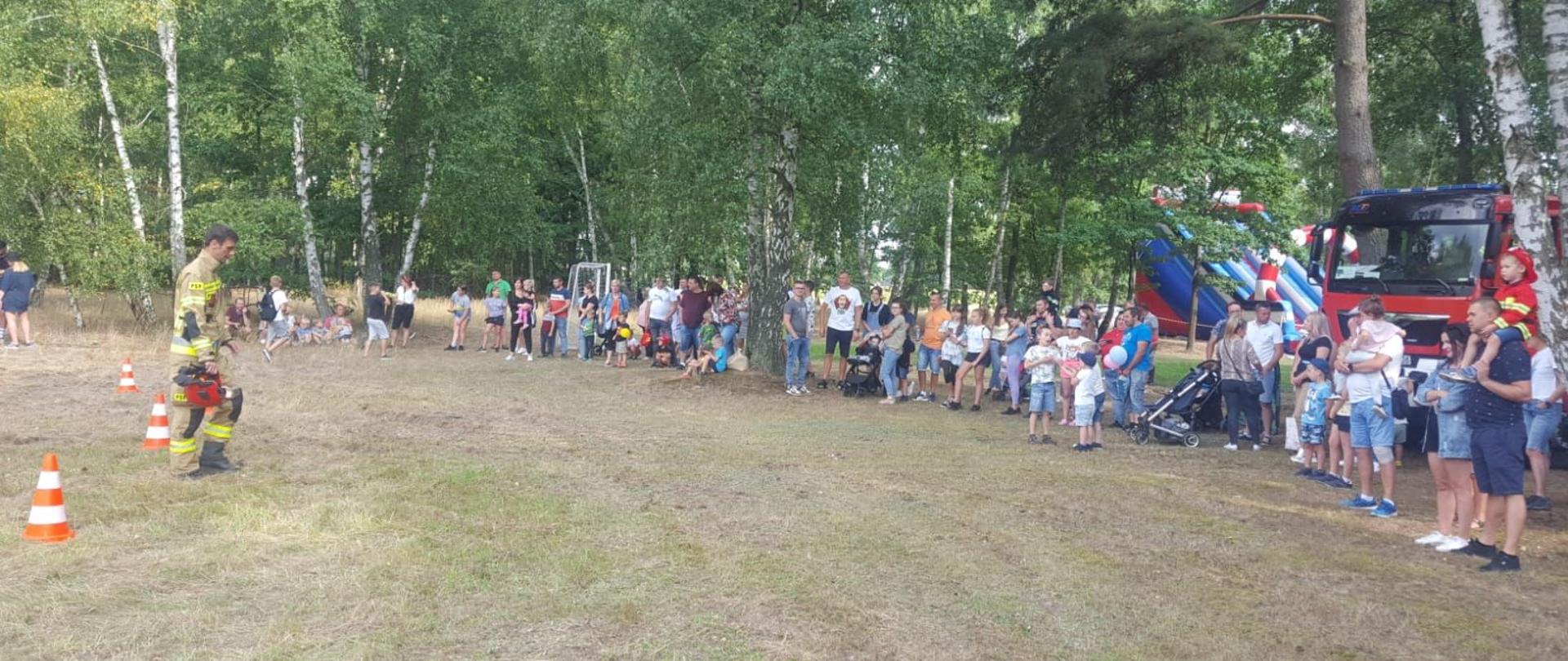 Dzień. Strażak w ubraniu specjalnym z hełmem w ręce stoi przed zebranymi uczestnikami festynu.