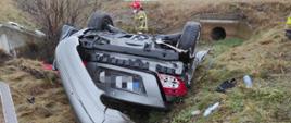 Zdjęcie przedstawia leżący na dachu samochód osobowy, przy którym znajduje się strażak.