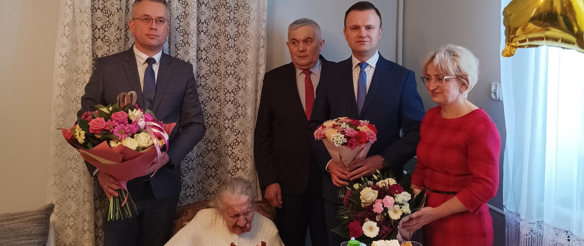 Jubilatka siedzi przy stole z tyłu trzej przedstawiciele lokalnych instytucji z bukietami kwiatów, trzech mężczyzn po lewej i kobieta po prawej