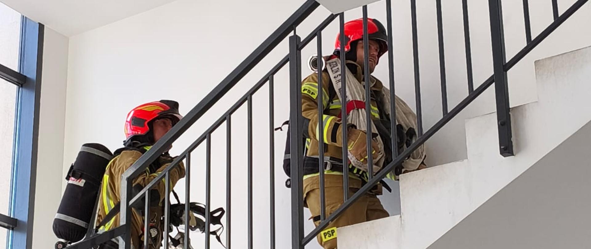 Strażacy w pełnym wyposażeniu wchodzą na górę po klatce schodowej.