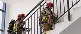 Strażacy w pełnym wyposażeniu wchodzą na górę po klatce schodowej.
