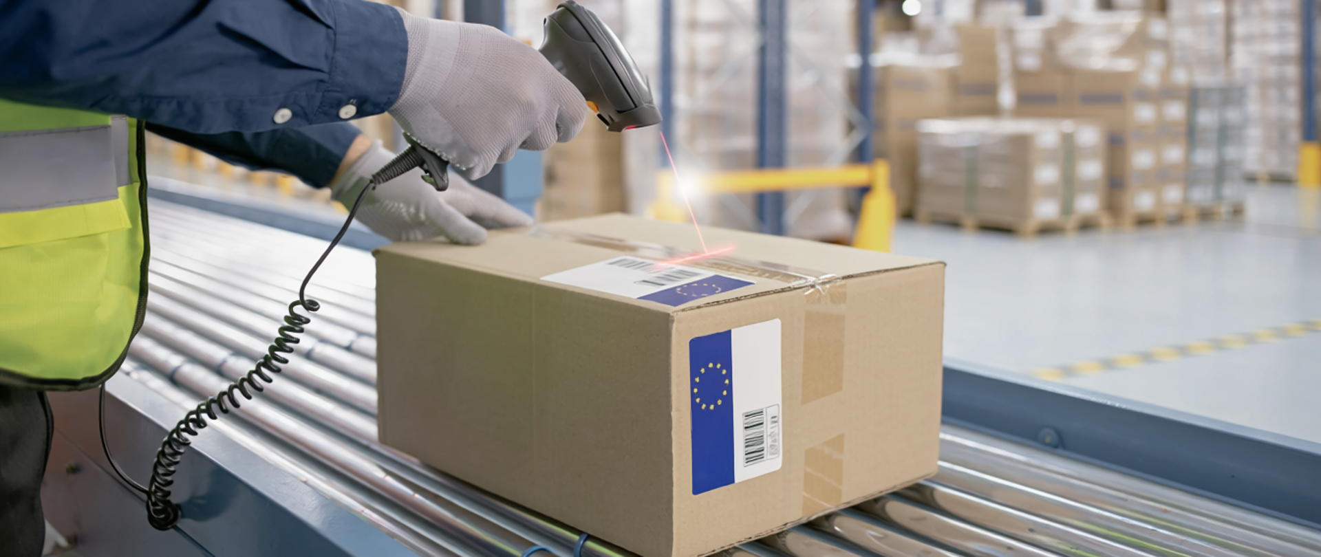 Pracownik magazynu skanuje kod kreskowy na kartonowej paczce za pomocą czytnika ręcznego. Paczka z etykietą i symbolem Unii Europejskiej leży na taśmie transportowej. W tle widać regały z towarem i inne zapakowane przesyłki.