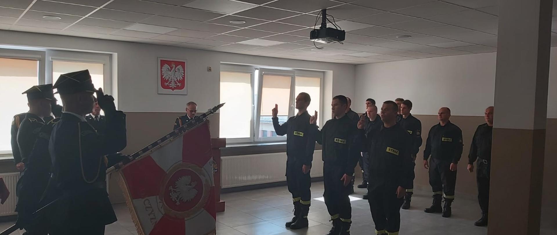 Po środku strażacy składają rotę ślubowania. Po lewej poczet sztandarowy oddaje honory. Z prawej strony stoi zmiana służbowa.