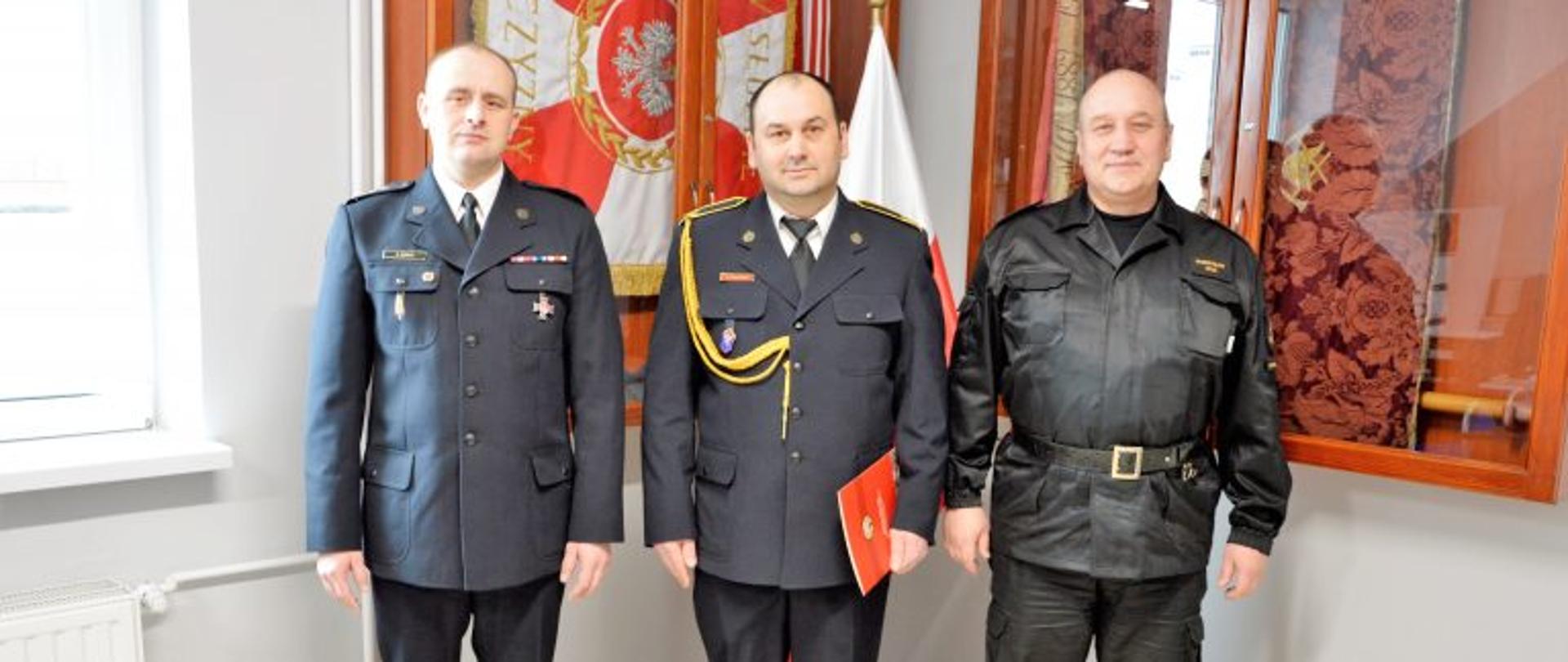 Na pamiątkowym zdjęciu Komendant Powiatowy wraz z zastępcą i Krzysztofem Orłowskim. Komendant oraz Krzysztof Orłowski w mundurach galowych. Zastępca w mundurze służbowym. W tle witryny ze sztandarem oraz biało czerwona flaga.
