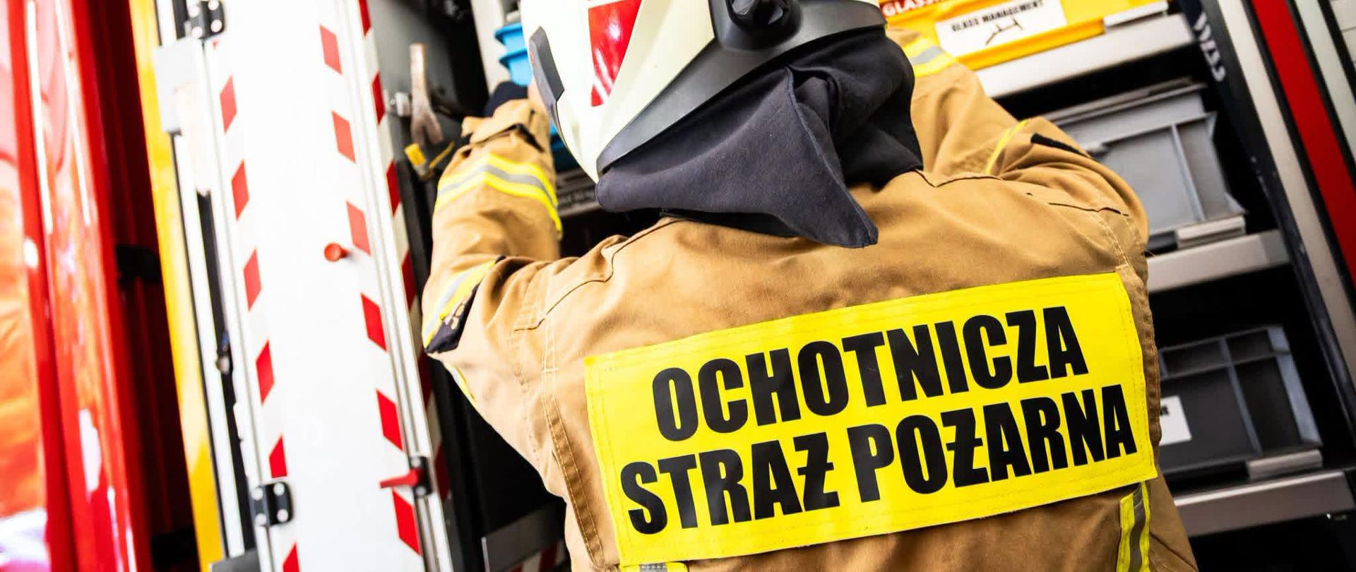 Strażak ochotnik w ubraniu ochronnym stoi tyłem do aparatu i sięga po sprzęt znajdujący się w otwartym przedziale wozu strażackiego. Na plecach ma żółtą naszywkę z dużym czarnym napisem „OCHOTNICZA STRAŻ POŻARNA”. Na białym hełmie widoczne są oznaczenia ratownika medycznego.