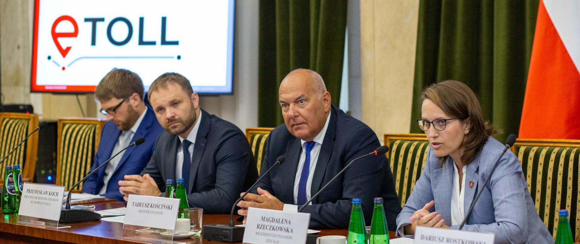Stół prezydialny przy którym siedzą minister finansów Tadeusz Kościński, szefowa KAS Magdalena Rzeczkowska, pełnomocnik ministra finansów ds. informatyzacji Przemysław Koch, w tle slajd prezentacji z napisem e-Toll.