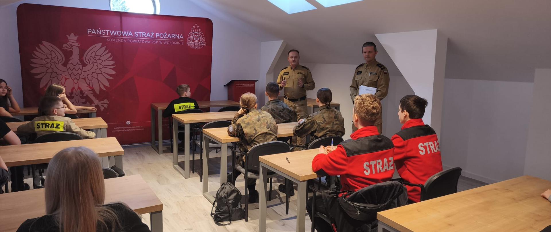 Strażak prowadzi zajęcia dla dzieci i młodzieży siedzących przy szkolnych ławkach w sali Komendy Państwowej Straży Pożarnej.