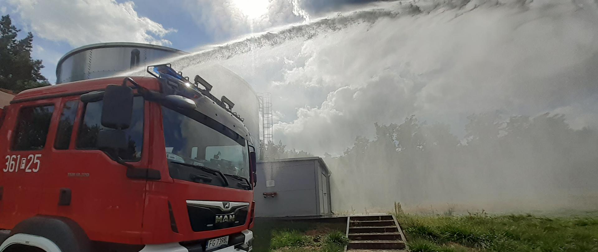 Zdjęcie przedstawia samochód pożarniczy na tle zbiornika przeciwpożarowego.
Z działka pojazdu pożarniczego podawany jest prąd wody.
