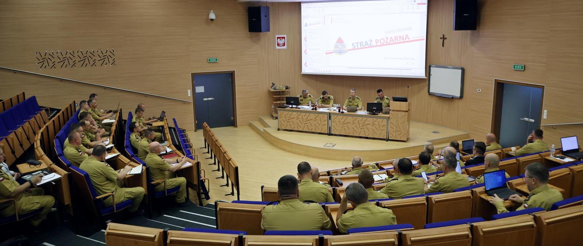 Widok z tyłu, na auli siedzą funkcjonariusze PSP podczas narady, w głębi na dole siedzący za stołem prezydialnym czterej oficerowie PSP