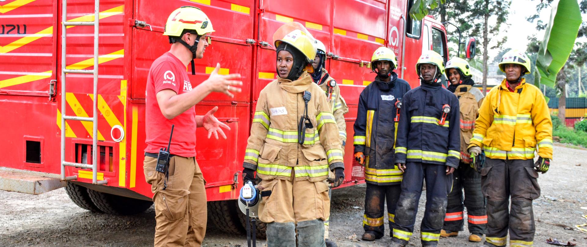 Obok samochodu strażackiego stoi grupa kobiet i mężczyzn i słucha instruktora, który rozmawia z jedną uczestniczką szkolenia