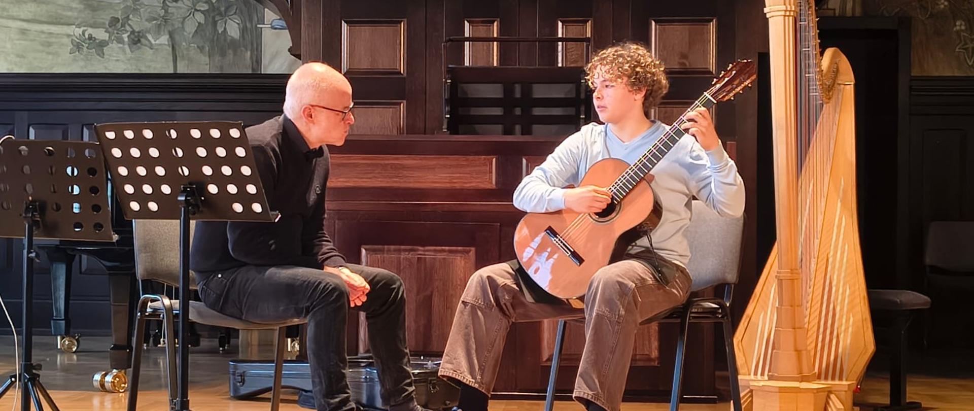 Kolorowe zdjęcie na którym znajduje się Maksymilian i prof. Andrzej Wilkus. Maksymilian siedzi na krześle i gra na gitarze, obok z lewej strony siedzi prof. A. Wilkus, który patrzy na Maksa i udziela mu cennych uwag.