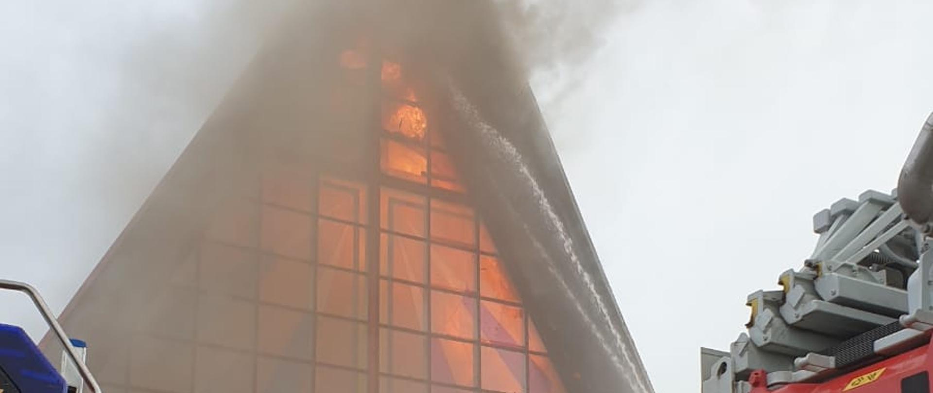 W centralnej części zdjęcia widok płonącego kościoła, płomienie wydostają się przez wybite w górnej części witraże. W dole po obu stronach stoją pojazdy pożarnicze i sylwetki strażaków rozwijających węże.
