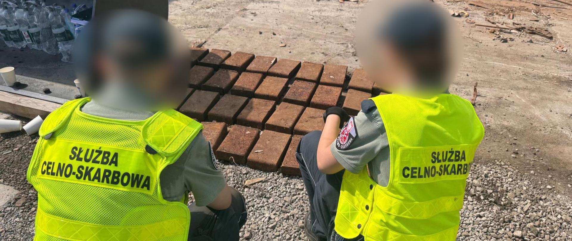 Funkcjonariusze Służby Celno-Skarbowej prowadzą oględziny prostokątnych sztabek metalu ułożonych w rzędach na placu przemysłowym.