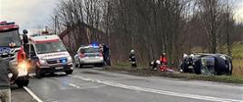 Zdjęcie wykonane na poboczu drogi w trakcie prowadzenia akcji ratowniczej. Po prawej stronie poza jezdnią samochód po dachowaniu, który leży na boku. W pobliżu strażacy oraz ratownik medyczny prowadzą działania. Po lewej stronie na drodze stoją pojazdy służb ratunkowych z włączonymi światłami błyskowymi niebieskimi, które blokują ruch na drodze. 