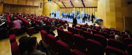 Perspektywa od tyłu sali widowiskowej. Widzowie siedzą w czerwonych fotelach, patrząc w stronę oświetlonej sceny, na której stoi grupa kilkunastu osób (uczestnicy i organizatorzy konkursu akordeonowego). Sala ma nowoczesny design z geometrycznymi panelami akustycznymi na suficie oraz drewnianym wykończeniem ścian.