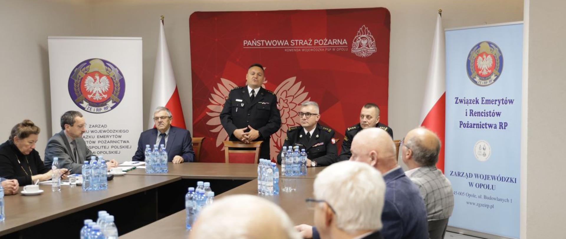 Spotkanie Seniorów Trzech Zarządów Oddziałów Wojewódzkich Związku Emerytów i Rencistów Pożarnictwa RP z Komendantami. Za stołem prezydialnym siedzą: Zastępca Dolnośląskiego Komendanta Wojewódzkiego PSP st. bryg. Piotr Grzyb oraz Zastępca Opolskiego Komendanta Wojewódzkiego PSP st. bryg. Arkadiusz Kuśmierski. W tle widoczny banner Państwowej Straży Pożarnej i Komendy Wojewódzkiej PSP w Opolu, dwa rollupy Związku Emerytów i Rencistów Pożarnictwa RP oraz dwie Flagi RP.
Na stojąco przemawia Opolski Komendant Wojewódzki PSP w Opolu st. bryg. Krzysztof Kędryk.