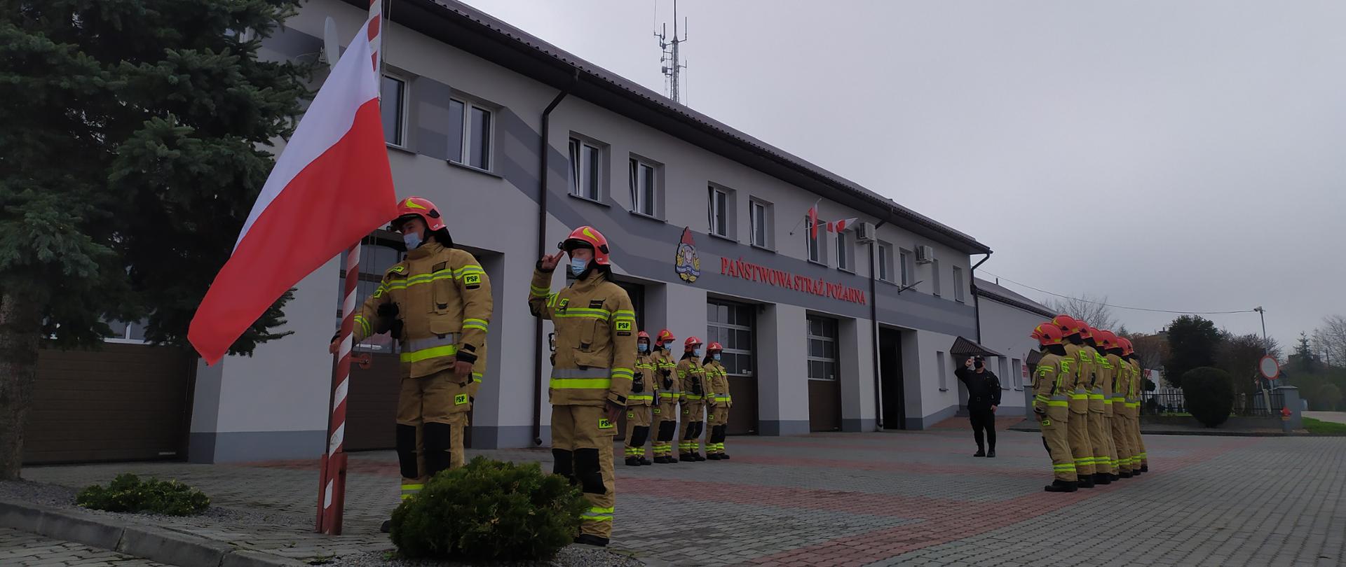 Zdjęcie przedstawia uroczystą zbiórkę pod budynkiem Komendy Powiatowej PSP w Lubaczowie. Po lewej stronie widoczni są strażacy w ubraniu specjalnym którzy podnoszą flagę państwową na maszt. W tle widoczne dwa szeregi strażaków w ubraniu specjalnym, a pomiędzy nimi, strażak w ubraniu dowódczo-sztabowym.